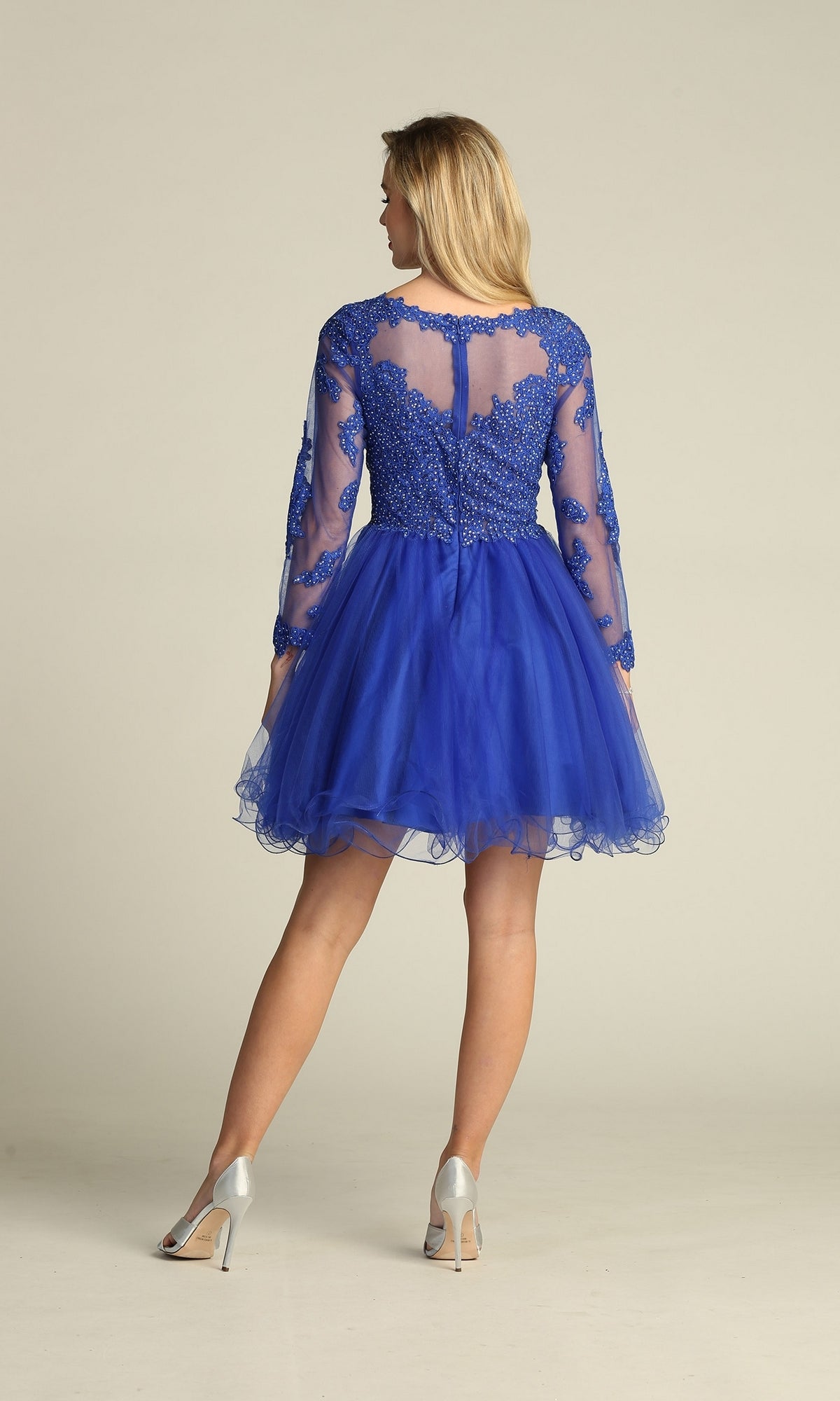 A woman stands facing away wearing the Beaded Long Sleeve Short Homecoming Dress DM4003 in royal blue, featuring sheer long sleeves, lace detailing, and a tulle skirt.